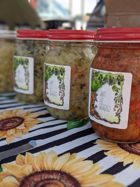 Kimchi and sauerkraut in jars that say "what got gathered" being sold at the farmers market.