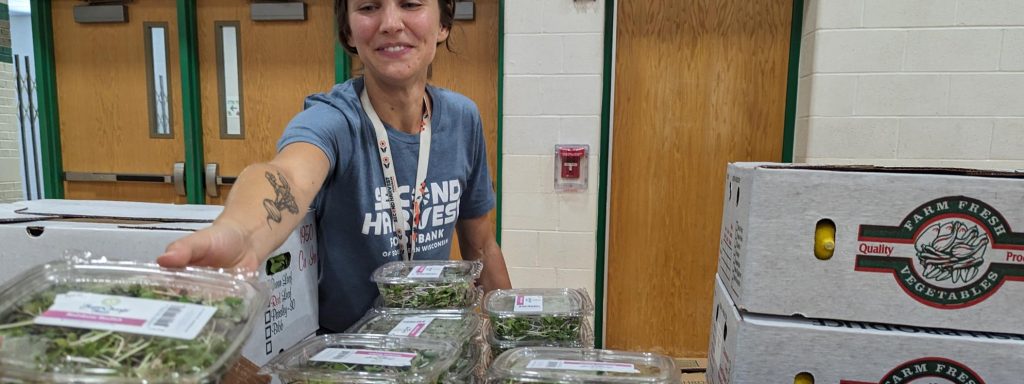 Jordyn at Beaver Dam WIC Family Festival. Woman wearing Second Harvest t-shirt reaches over to hand someone a box of green.